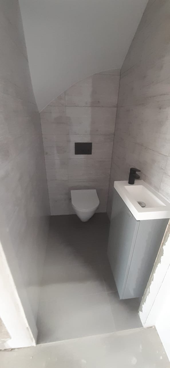 Mała łazienka w odcieniach szarości z podwieszaną toaletą i umywalką z szafką, czarna bateria umywalkowa i przycisk spłuczki.