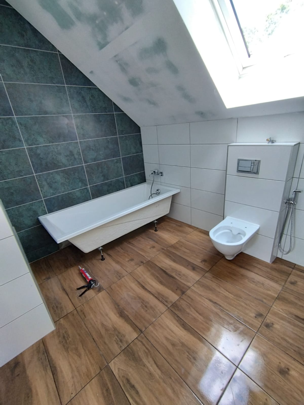 Ukończona łazienka na poddaszu ze skośnym sufitem, z wanną na nóżkach, podwieszaną toaletą i drewnopodobnymi płytkami podłogowymi, widoczne narzędzia do silikonowania.