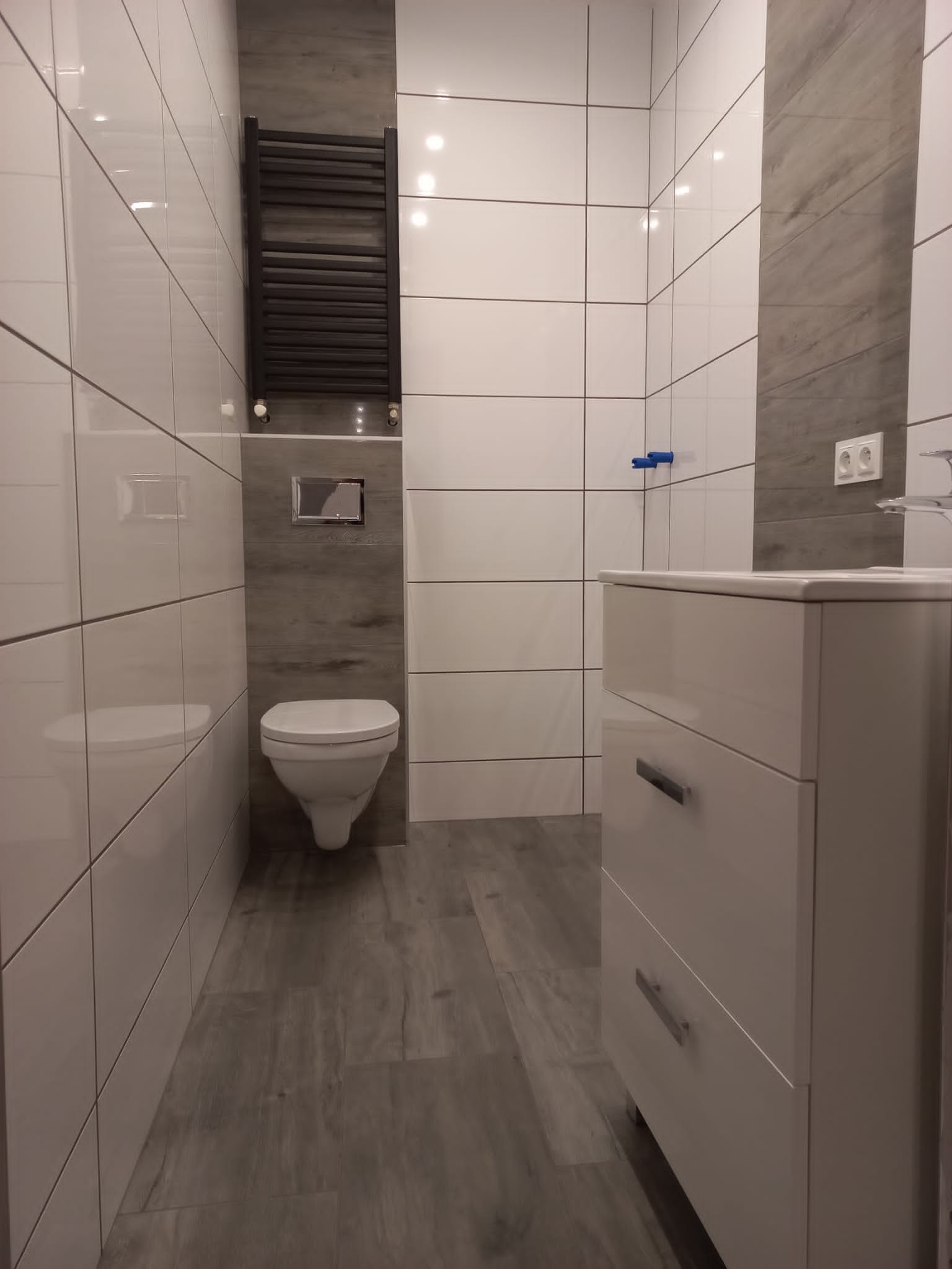 Nowoczesna łazienka z białymi, błyszczącymi płytkami, grzejnikiem drabinkowym, podwieszaną toaletą i szafką umywalkową z szufladami.