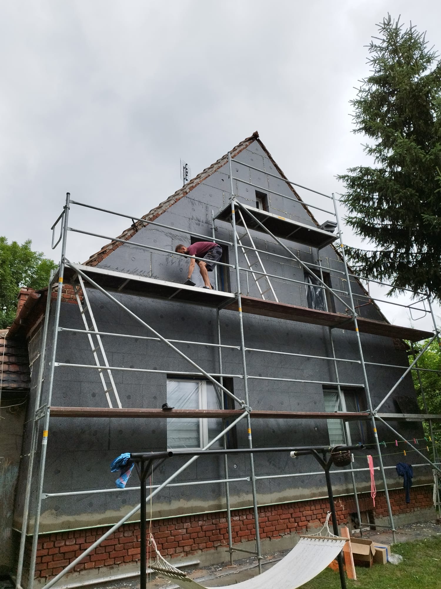 Pracownik na rusztowaniu mocuje szare płyty izolacyjne do ściany domu z ceglaną podmurówką, widok z dołu na budynek w trakcie ocieplania.