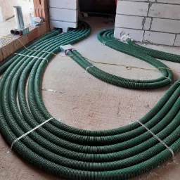 Zielone, karbowane rury wentylacyjne ułożone równolegle na podłodze w surowym stanie, przymocowane metalowymi opaskami, prowadzące do skrzynki rozdzielczej i otworów w ścianie z pustaków.