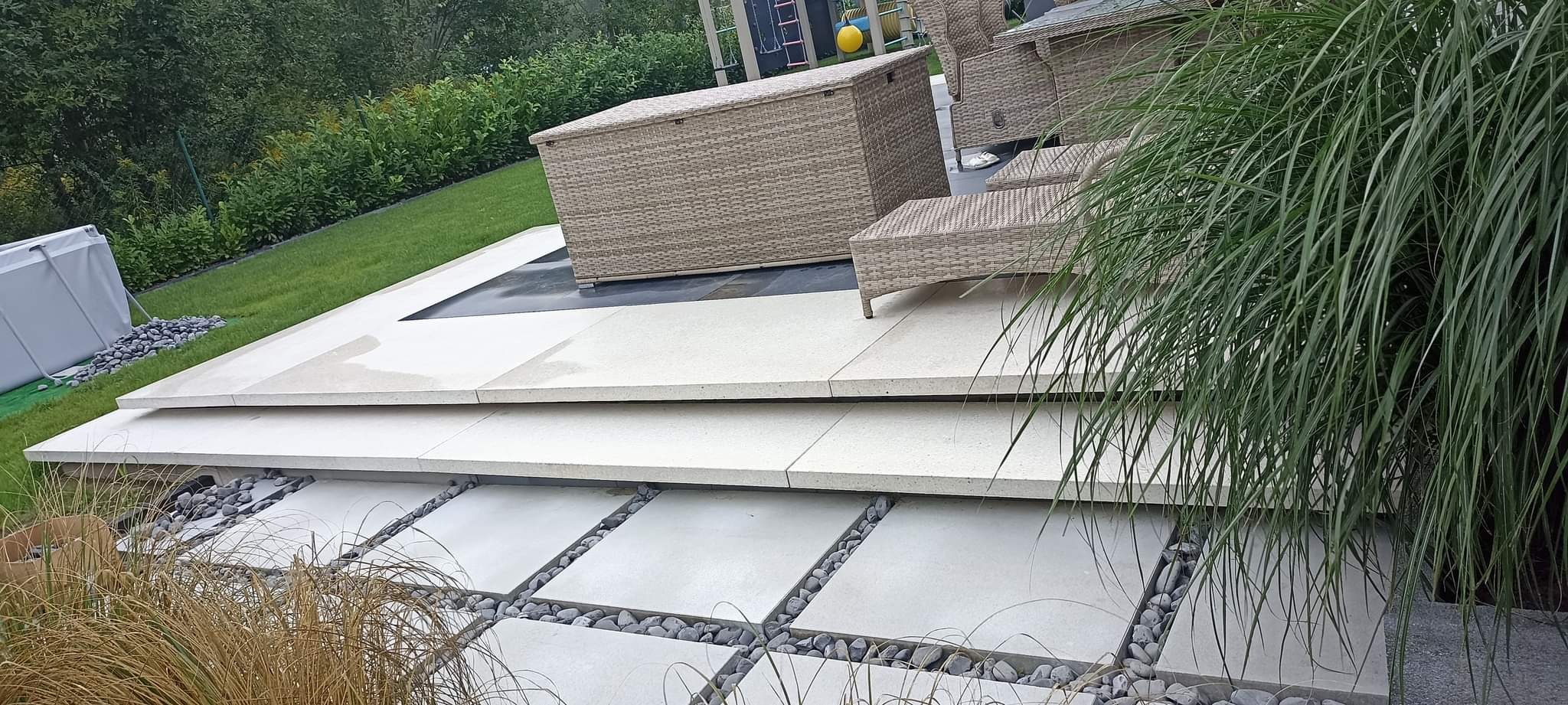 Wielopoziomowy taras z jasnych płyt betonowych z dekoracyjnymi kamieniami, uzupełniony meblami ogrodowymi z rattanu i elementami zieleni.