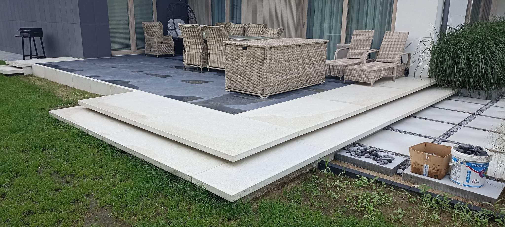 Nowoczesny taras z ciemną nawierzchnią i jasnymi schodami z płyt. Widoczne meble ogrodowe z rattanu i roślinność ozdobna. Na pierwszym planie materiały budowlane: kamienie i wiadro z zaprawą.