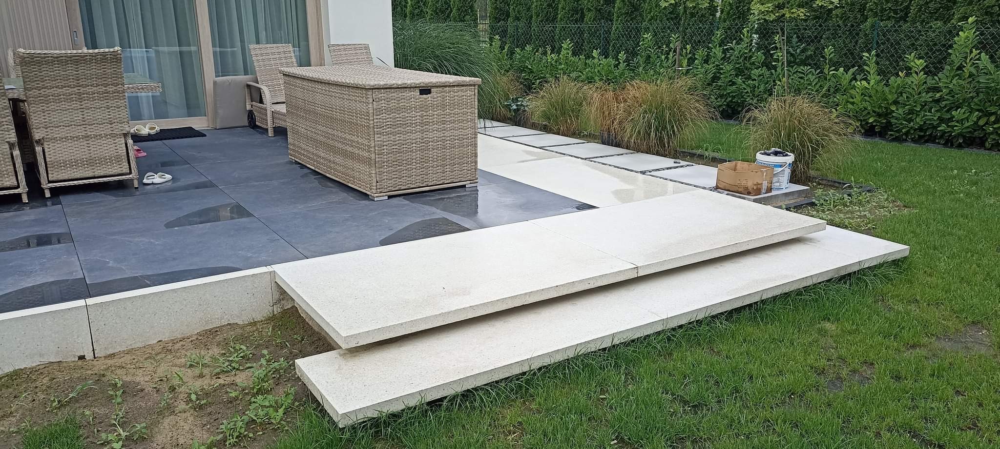 Dwa stopnie schodowe z jasnego kamienia, ułożone na trawie, prowadzące na taras z ciemnych płyt. W tle meble ogrodowe z rattanu i dekoracyjna roślinność.