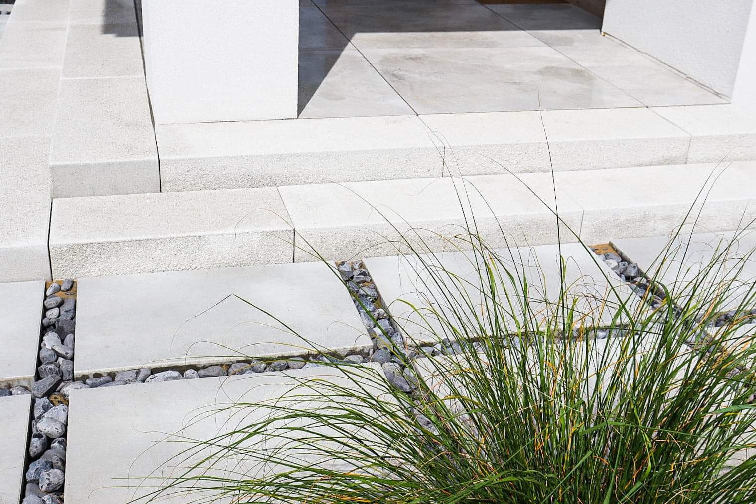 Jasnoszare płyty tarasowe z betonowymi schodami, oddzielone dekoracyjnymi pasami z kamyków, z zieloną trawą na pierwszym planie.