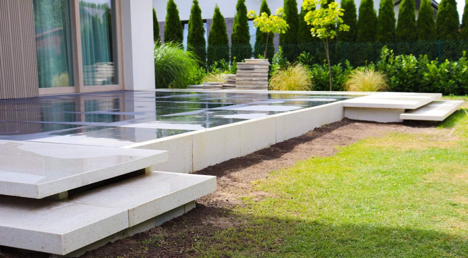 Nowoczesny taras z błyszczących płyt w szachownicę, z niską ścianką i schodami z jasnego kamienia, otoczony zielenią i tujami.