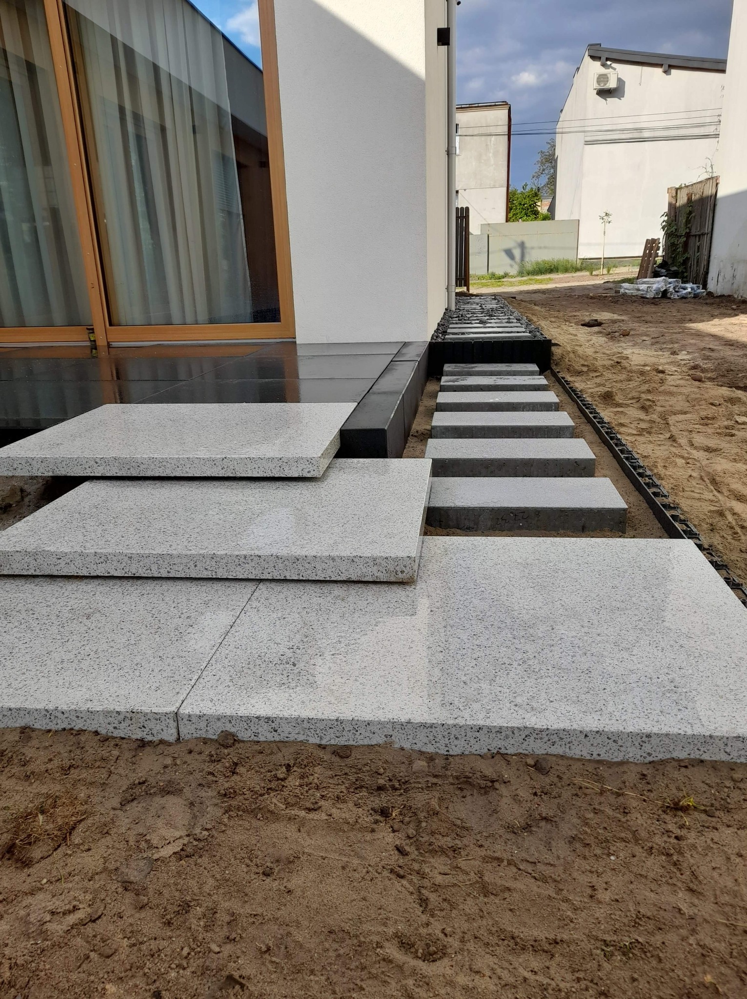 Nowoczesne schody zewnętrzne z jasnoszarego kamienia prowadzące do domu, z widocznymi płytami chodnikowymi w ogrodzie, w tle biały budynek i pochmurne niebo.