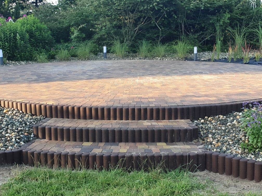 Okrągły taras z kostki brukowej w odcieniach brązu i beżu, z dwoma stopniami obłożonymi kostką palisadową, otoczony ozdobnymi kamieniami i zielenią. W tle wysokie trawy i krzewy.