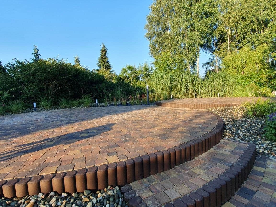 Okrągły plac wyłożony kostką brukową w odcieniach brązu i czerwieni, z obrzeżem z ciemnobrązowych, walcowatych elementów. Dwa stopnie prowadzą na wyższy poziom placu. W tle bujna roślinność, w tym...