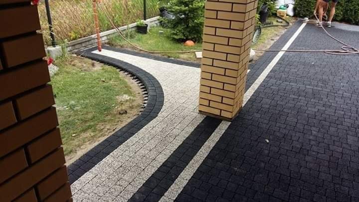 Ułożona kostka brukowa w dwóch kolorach: czarnym i jasnoszarym, tworząca ścieżkę i podjazd. Ścieżka wykończona obrzeżem z kostki. Na podjeździe widoczna biała linia oddzielająca pasy ruchu. W tle...