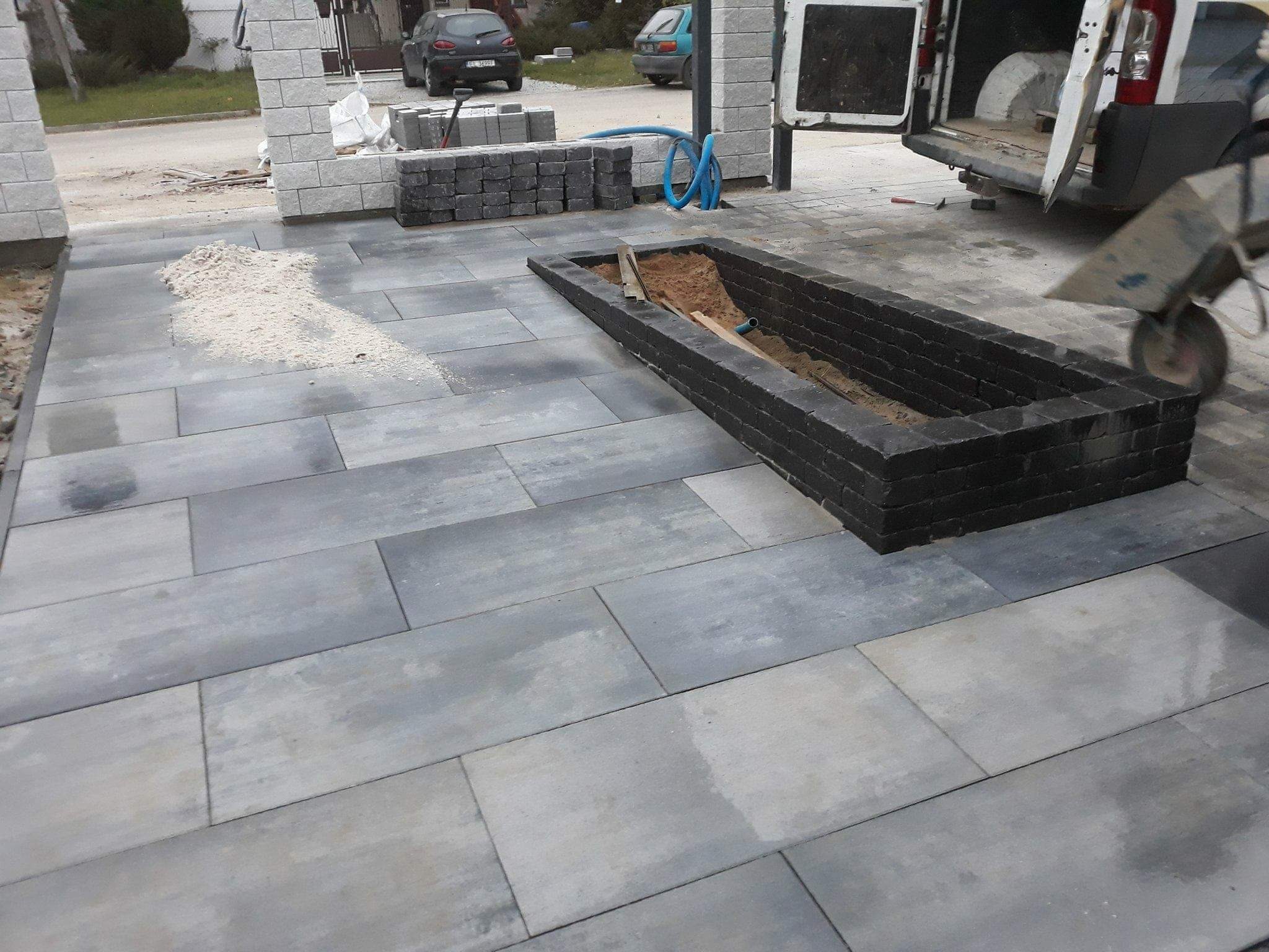 Niedokończona nawierzchnia z szarych płyt betonowych z przygotowanym obramowaniem z ciemnej kostki na rabatę kwiatową wypełnioną piaskiem, widoczny wózek transportowy i samochód dostawczy w tle.
