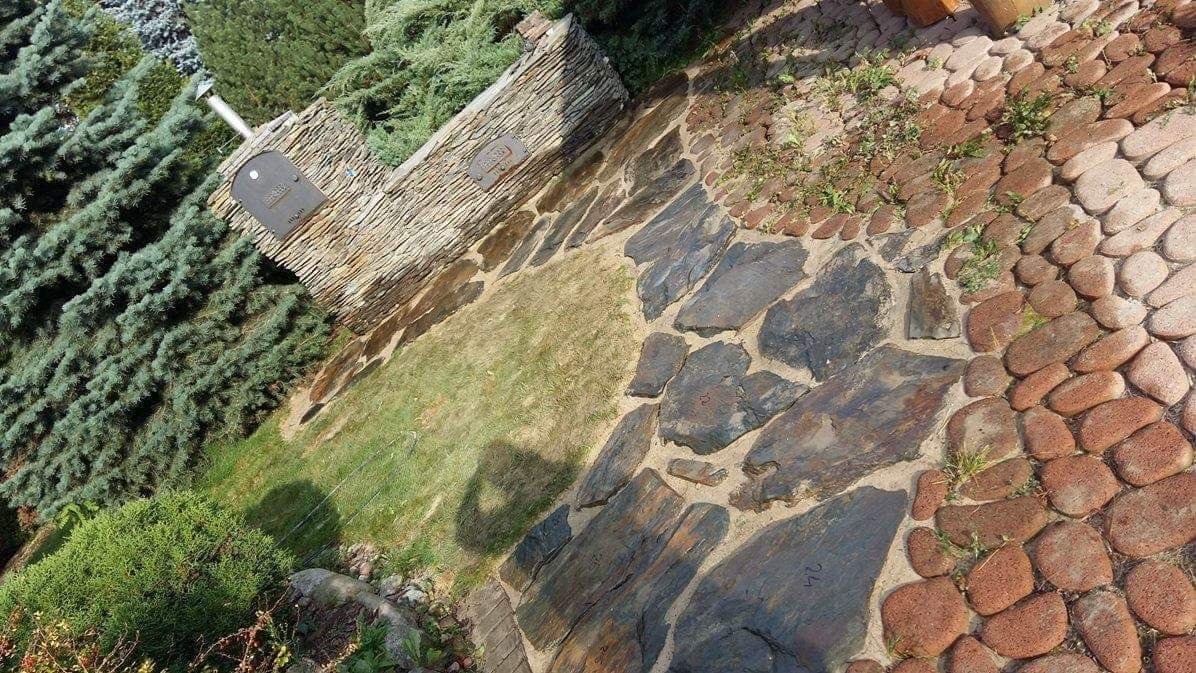 Ogród z ścieżką z nieregularnych kamieni w odcieniach szarości i brązu, obok ściana z kamienia łupanego i skrzynka pocztowa, trawa i zieleń w tle.