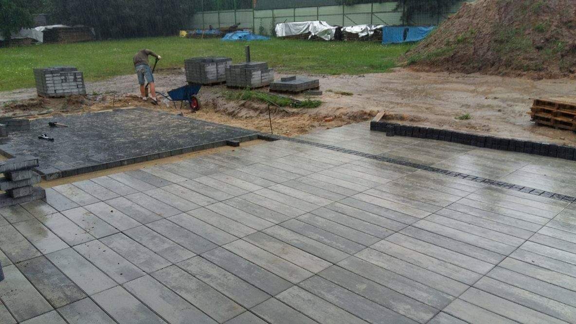 Montaż szarej kostki brukowej w deszczowy dzień, widoczny pracownik z łopatą i taczka, ułożona część nawierzchni z liniowym odprowadzeniem wody, palety kostki w tle.