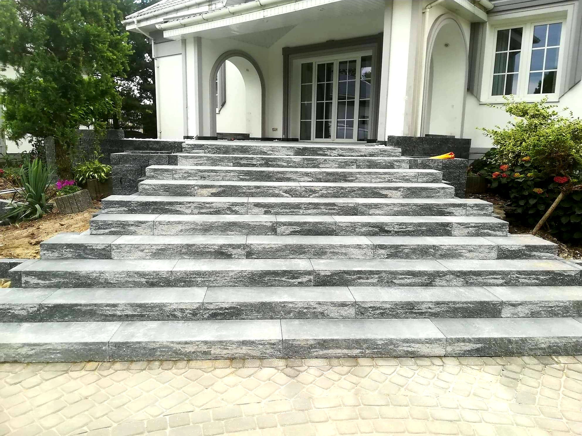 Kamienne schody prowadzące do wejścia domu, wykonane z szarych bloków o nieregularnej fakturze, z kostką brukową u podstawy i zielenią po bokach.