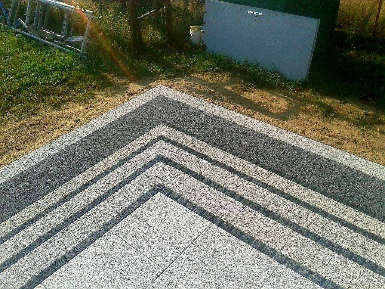 Schody zewnętrzne z jasnoszarej kostki granitowej, ułożone w warstwowy sposób, z widocznym piaskowym podłożem i drabiną w tle.