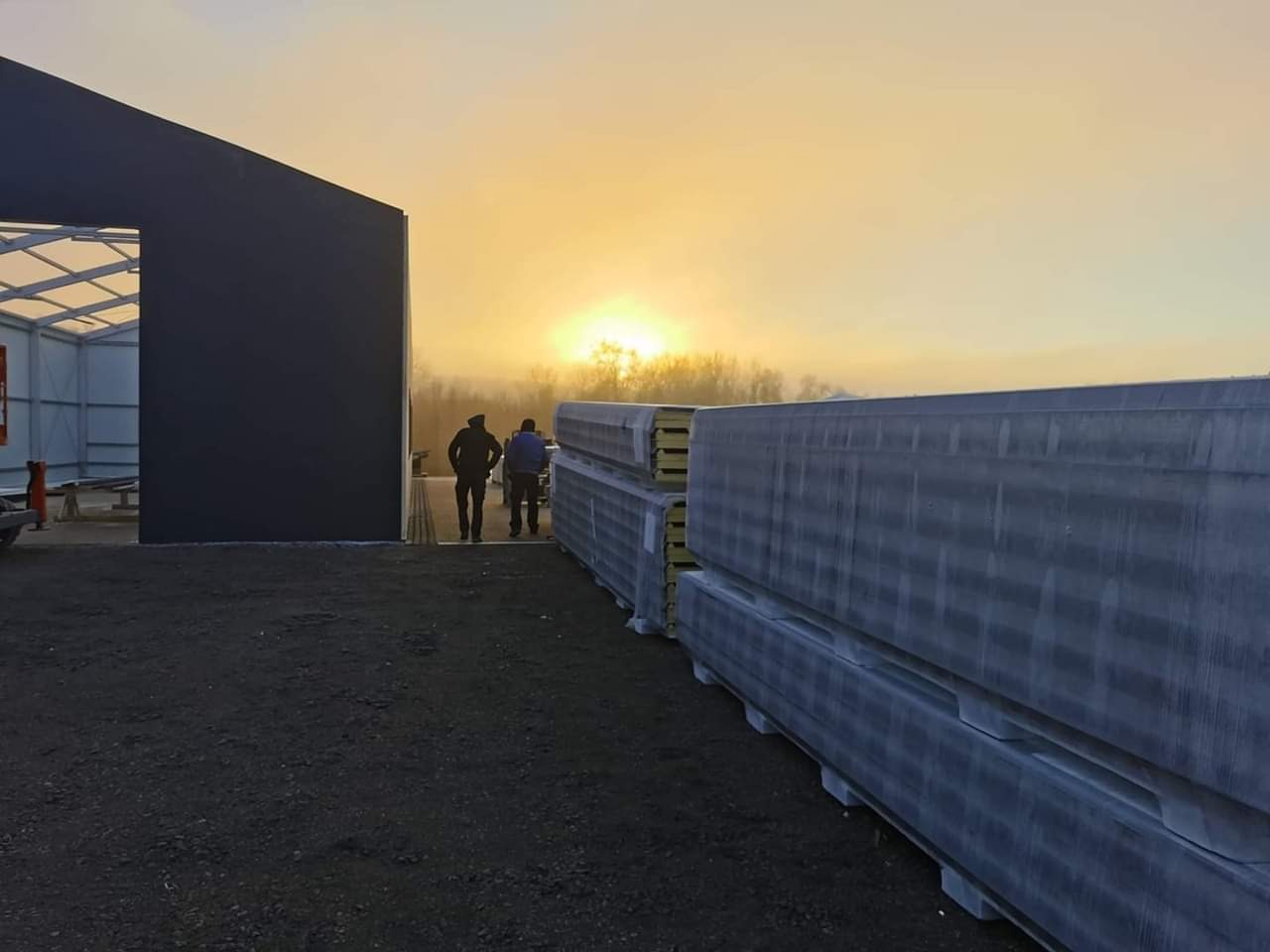 Wczesny poranek na placu budowy: szkielet hali z czarną elewacją, palety z panelami warstwowymi i sylwetki dwóch pracowników na tle wschodzącego słońca.