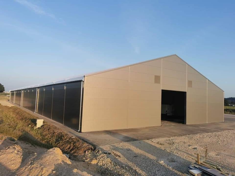 Gotowa hala magazynowa z beżowych paneli z czarnymi przesuwnymi bramami na betonowej wylewce, teren wokół w trakcie prac ziemnych.