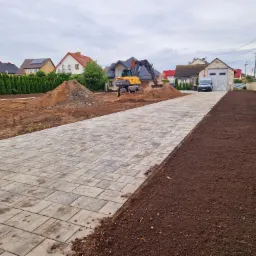 Żółta koparka Mecalac na placu budowy, wyrównuje teren obok ułożonej kostki brukowej, w tle domy jednorodzinne.