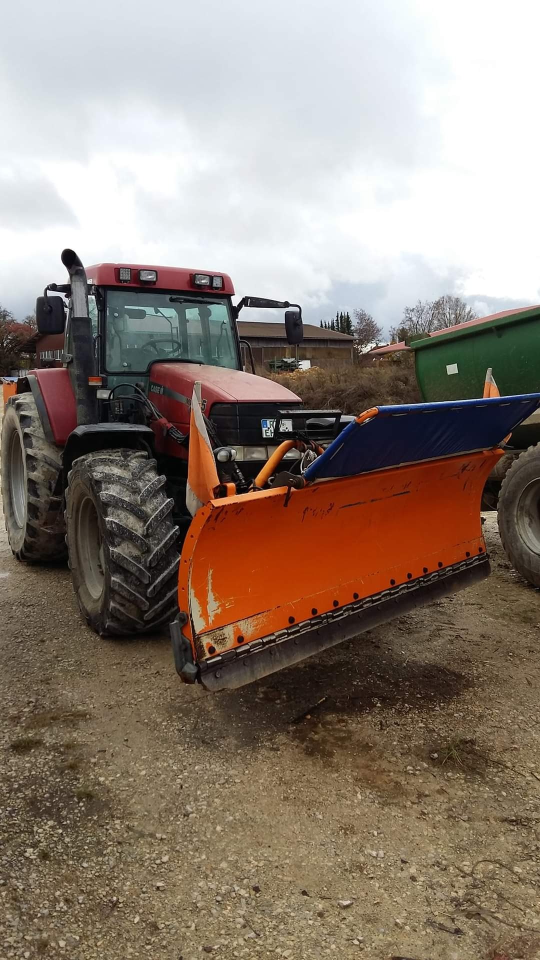 Czerwony traktor Case IH z pomarańczowym pługiem do odśnieżania, stojący na żwirowym podłożu pod pochmurnym niebem; widoczny fragment przyczepy w tle.