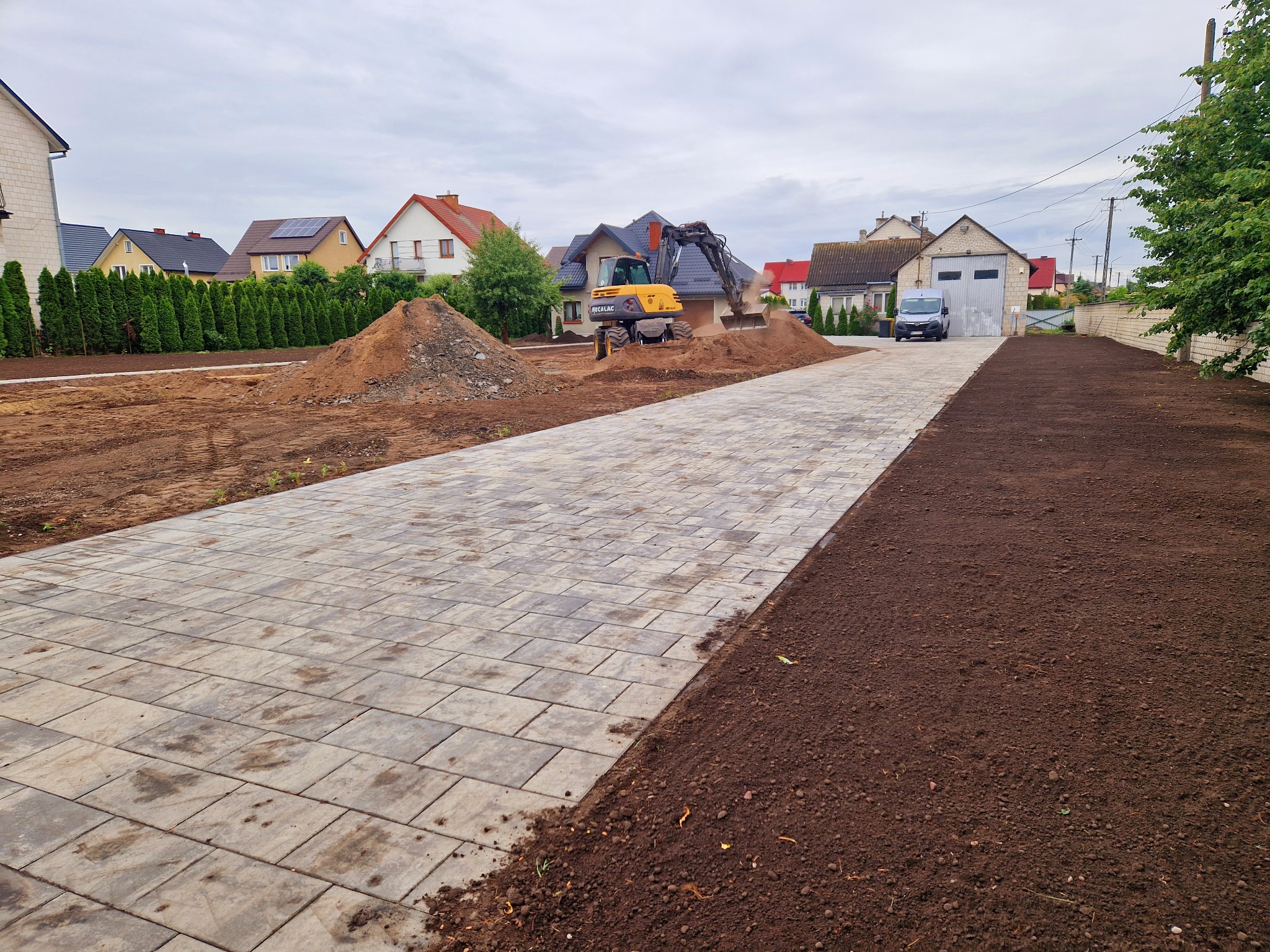 Żółta koparka Mecalac na placu budowy, wyrównuje teren obok ułożonej kostki brukowej, w tle domy jednorodzinne.