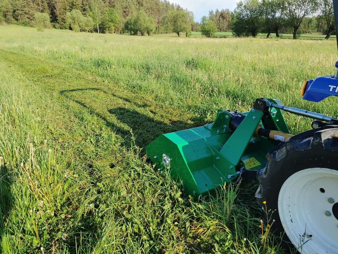 Zielony traktor z kosiarką bijakową w trakcie koszenia wysokiej trawy na łące, widoczny ślad po koszeniu.