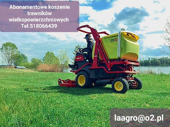 Czerwona kosiarka samojezdna z zielonym zbiornikiem tnąca trawę na rozległym, zielonym terenie w pobliżu jeziora, operator w słuchawkach ochronnych.