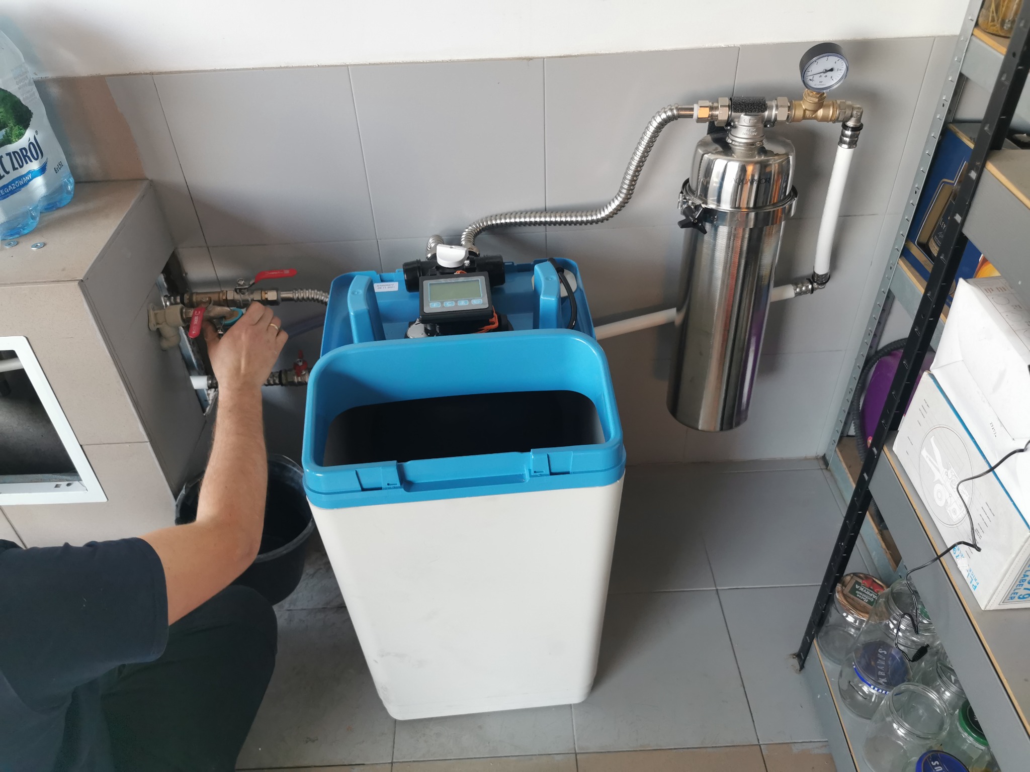 Instalacja zmiękczacza wody Aquaphor: hydraulik z pierścieniem na palcu reguluje zawór dopływu wody, widoczny srebrny filtr ciśnieniowy z manometrem oraz zbiornik na sól z niebieską pokrywą.