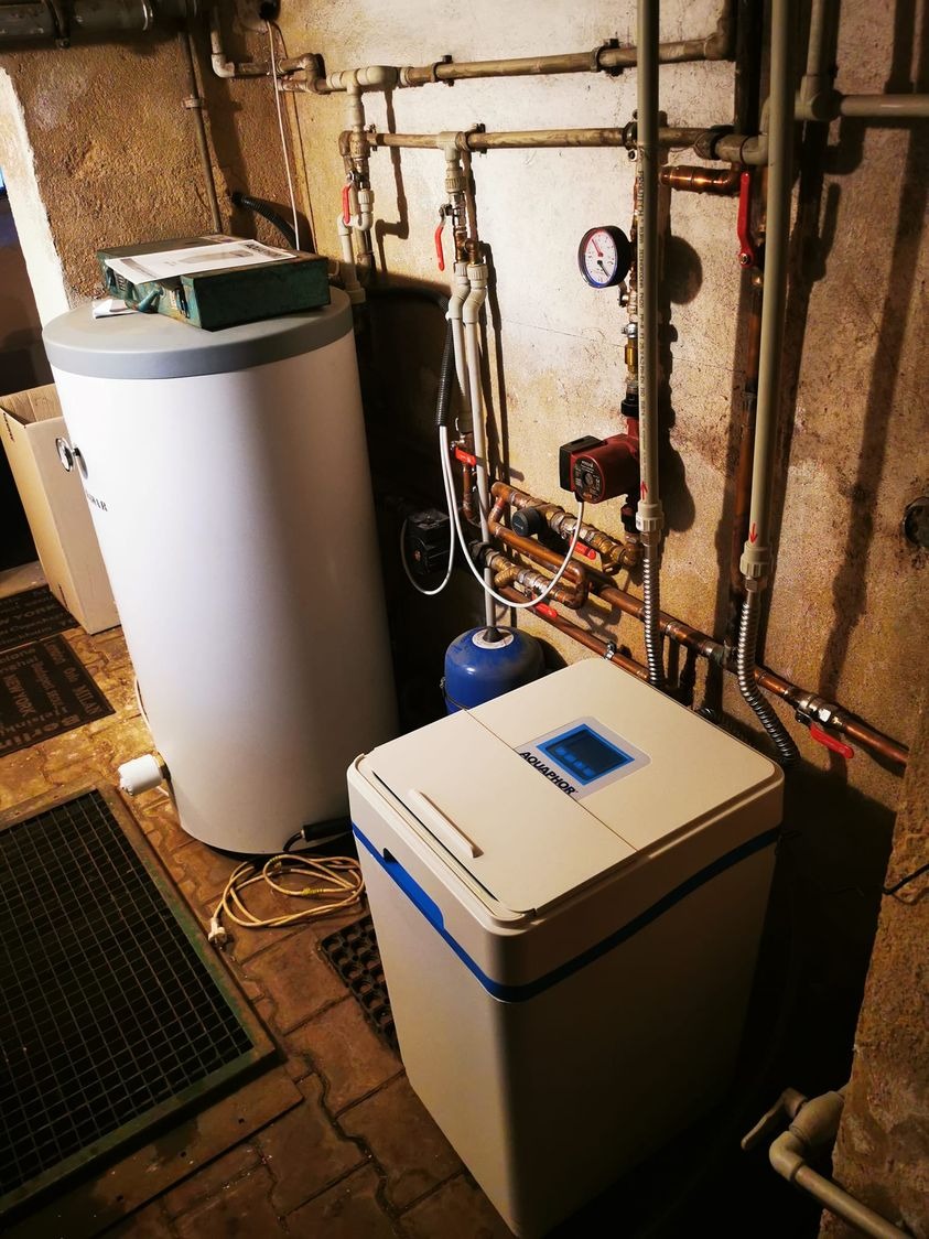 W piwnicy zestaw urządzeń: biały zbiornik na wodę z zielonym pudełkiem na górze, obok zmiękczacz wody Aquaphor z niebieskimi akcentami, w tle instalacja rur miedzianych i zaworów na surowej ścianie.
