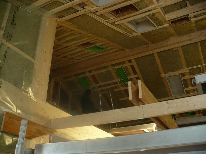 Wnętrze poddasza w trakcie izolacji: widoczne drewniane belki konstrukcyjne, wełna mineralna między krokwiami, folia paroizolacyjna na ścianie szczytowej oraz fragment drabiny aluminiowej.