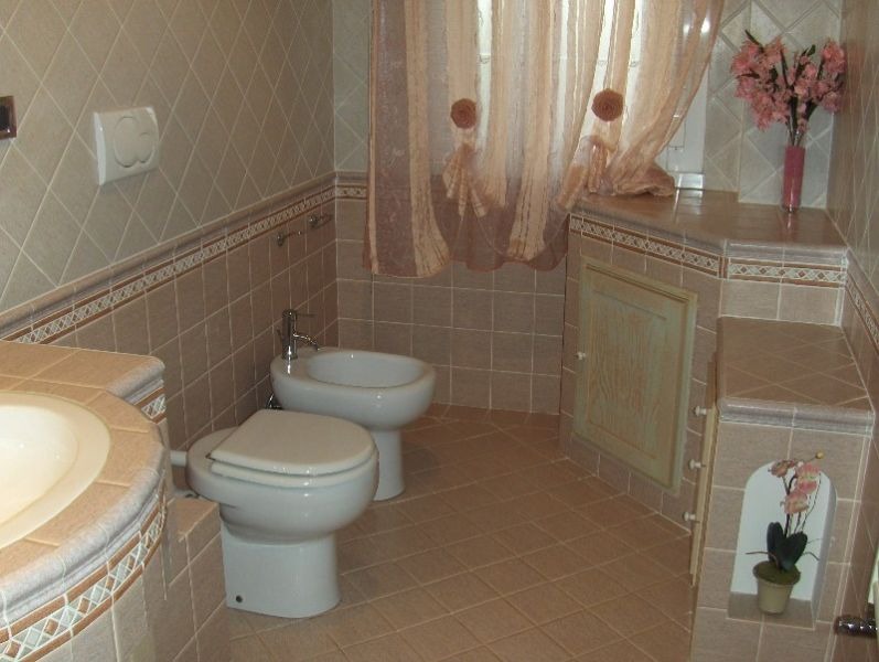 Beżowa łazienka z kwadratowymi płytkami ułożonymi w karo, białą armaturą (toaleta i bidet), zasłoną w kwiaty i dekoracyjną wnęką z kwiatami.