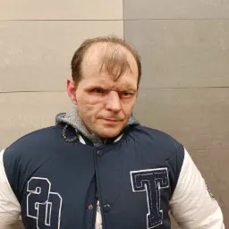 Mężczyzna w kurtce typu bejsbolówka z literą 'T' i liczbą '20' na tle szarej ściany z płytek.