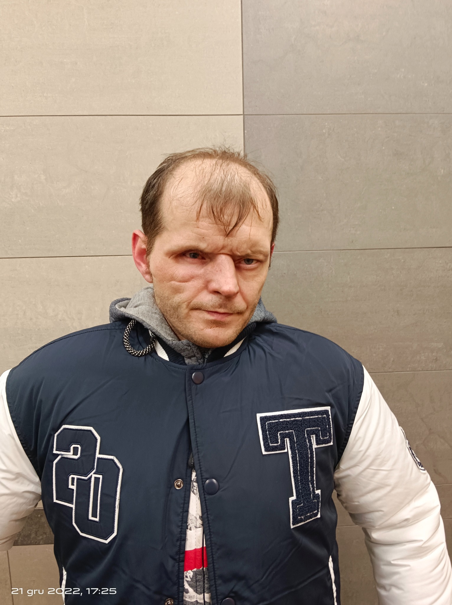 Mężczyzna w kurtce typu bejsbolówka z literą 'T' i liczbą '20' na tle szarej ściany z płytek.