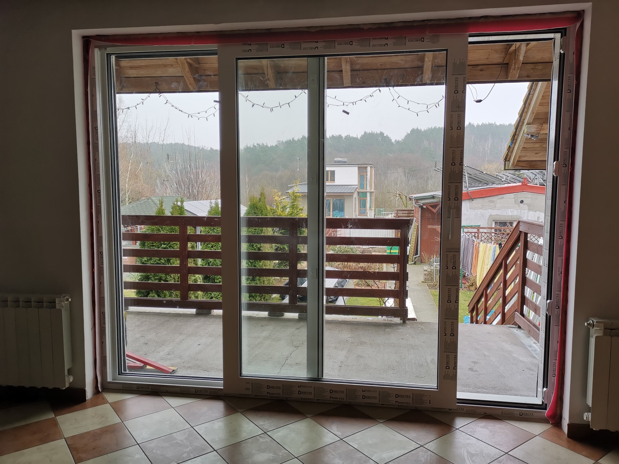 Białe, przesuwne drzwi balkonowe PCV z widokiem na zewnątrz, częściowo otwarte. Widoczny fragment wnętrza z grzejnikiem i wzorzystą podłogą. Na zewnątrz balkon i zabudowa.