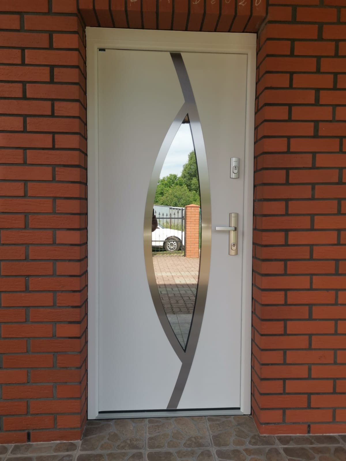 Białe drzwi zewnętrzne z dekoracyjnym, owalnym przeszkleniem i srebrnymi akcentami, wmurowane w czerwoną cegłę. Widok na podjazd i zieleń przez szybę.