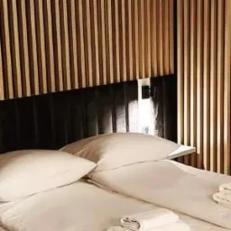 Minimalistyczna sypialnia z łóżkiem z białymi poduszkami i pościelą, wezgłowie z ciemnej tapicerki z pionowymi, drewnianymi listwami, oświetlenie punktowe.