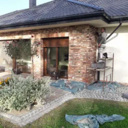 Pracownik na rusztowaniu, poprawiający fugi w ceglanej elewacji domu jednorodzinnego z ciemną dachówką, widoczne zabezpieczenia na ziemi.