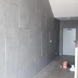 Korytarz wykończony szarymi płytkami imitującymi beton na ścianach i podłodze, z widocznym opakowaniem kleju do płytek Flex Active na podłodze.