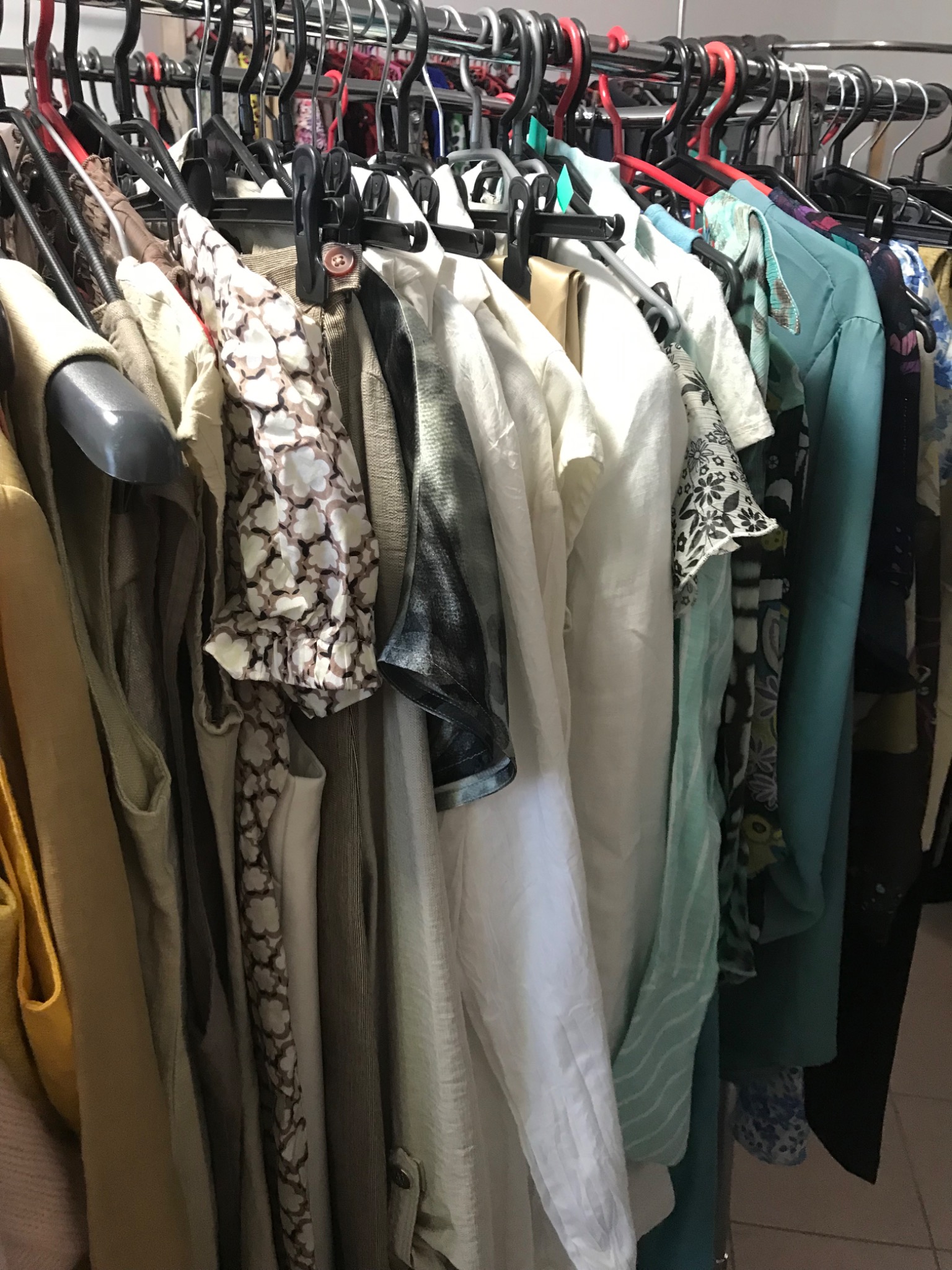 Rząd wieszaków z różnorodną odzieżą, głównie w jasnych, stonowanych kolorach, widoczne wzory kwiatowe i geometryczne, niektóre ubrania z krótkim rękawem, na jednym z wieszaków widoczny brązowy guzik.