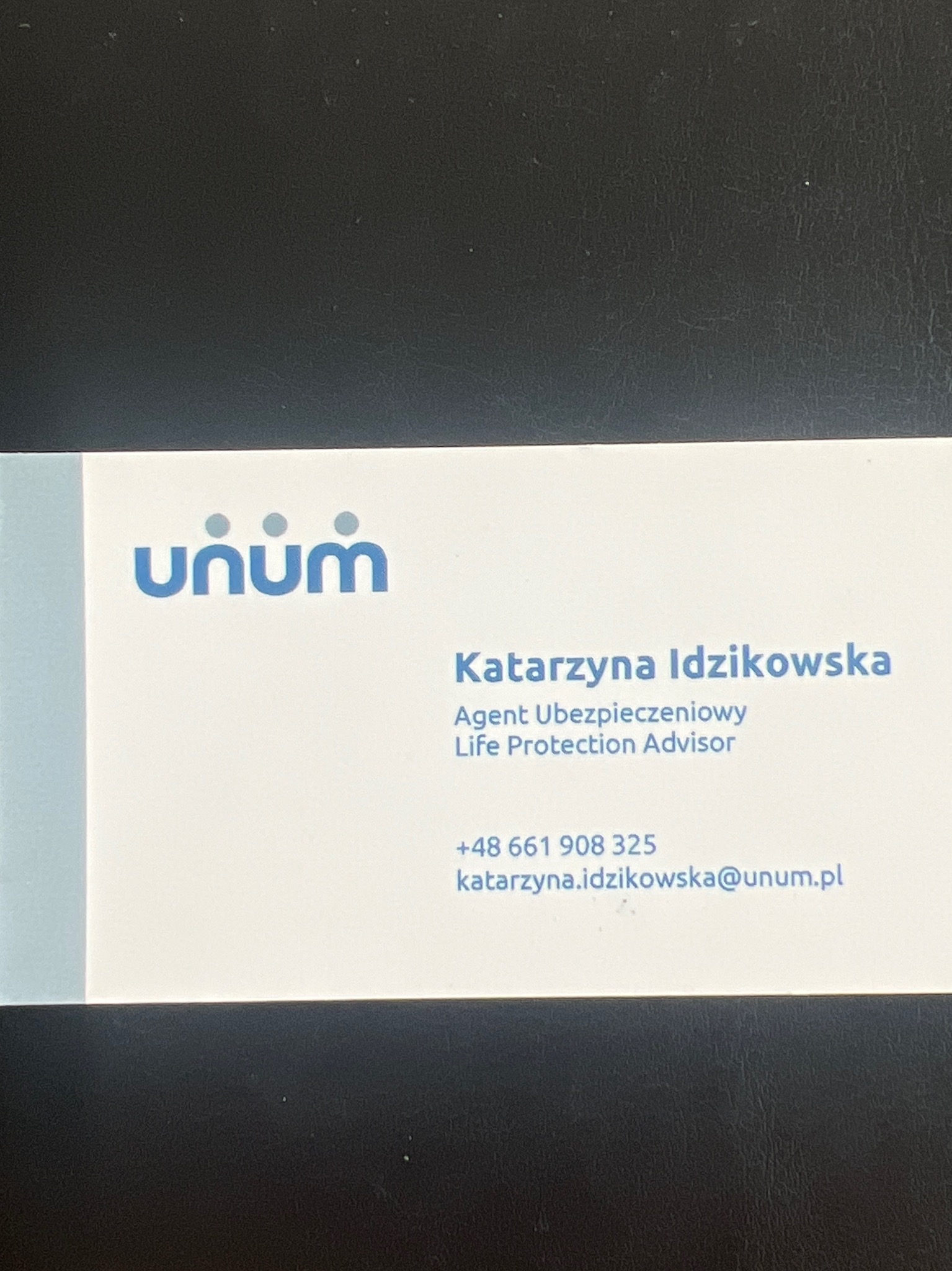 Wizytówka agenta ubezpieczeniowego Katarzyny Idzikowskiej z logo Unum, numerem telefonu i adresem email, leżąca na ciemnym tle.
