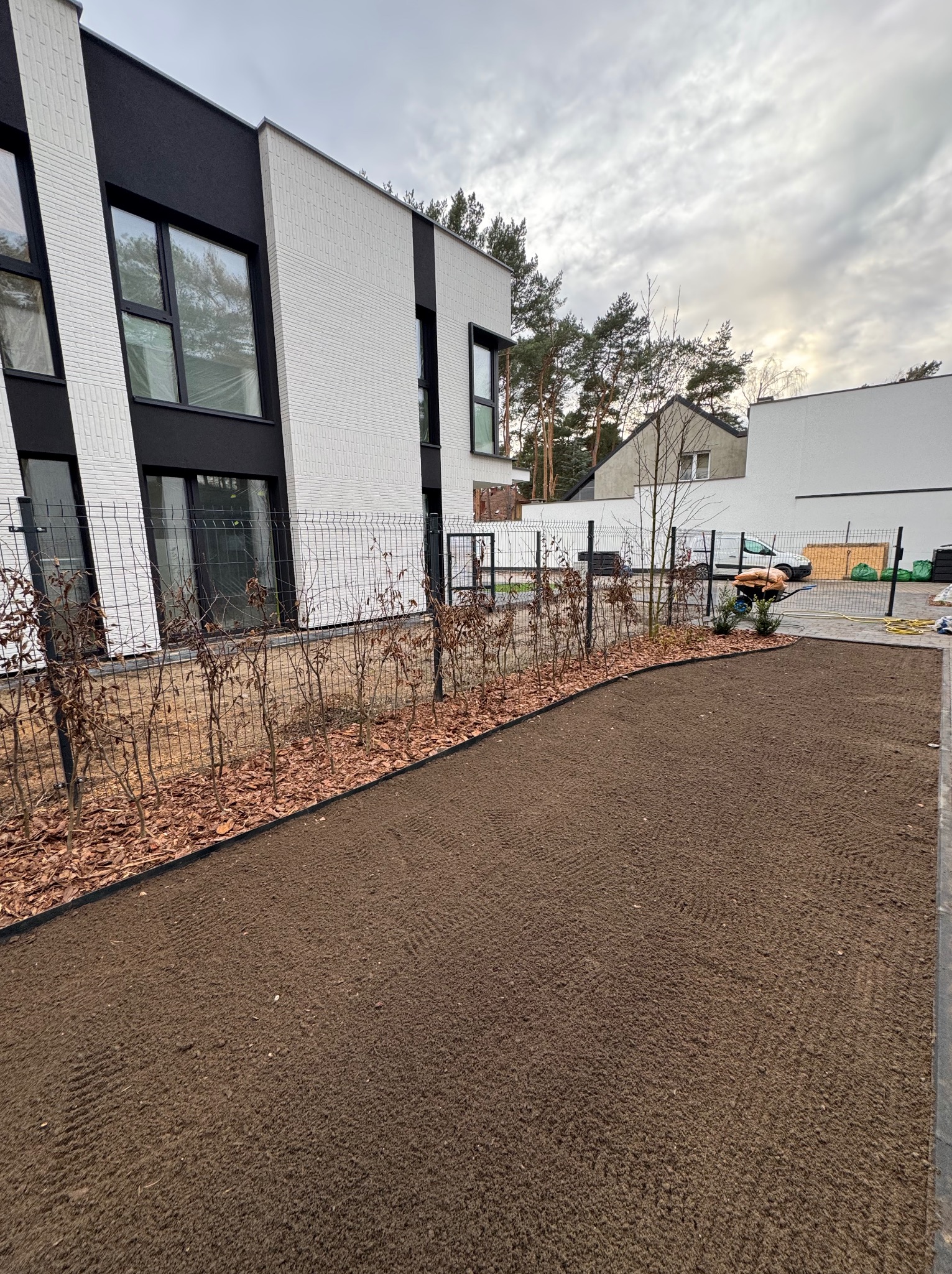 Przygotowany teren pod trawnik z rolki, obrzeża rabat, młode krzewy wzdłuż ogrodzenia na tle nowoczesnego domu z białej cegły i szarego nieba.