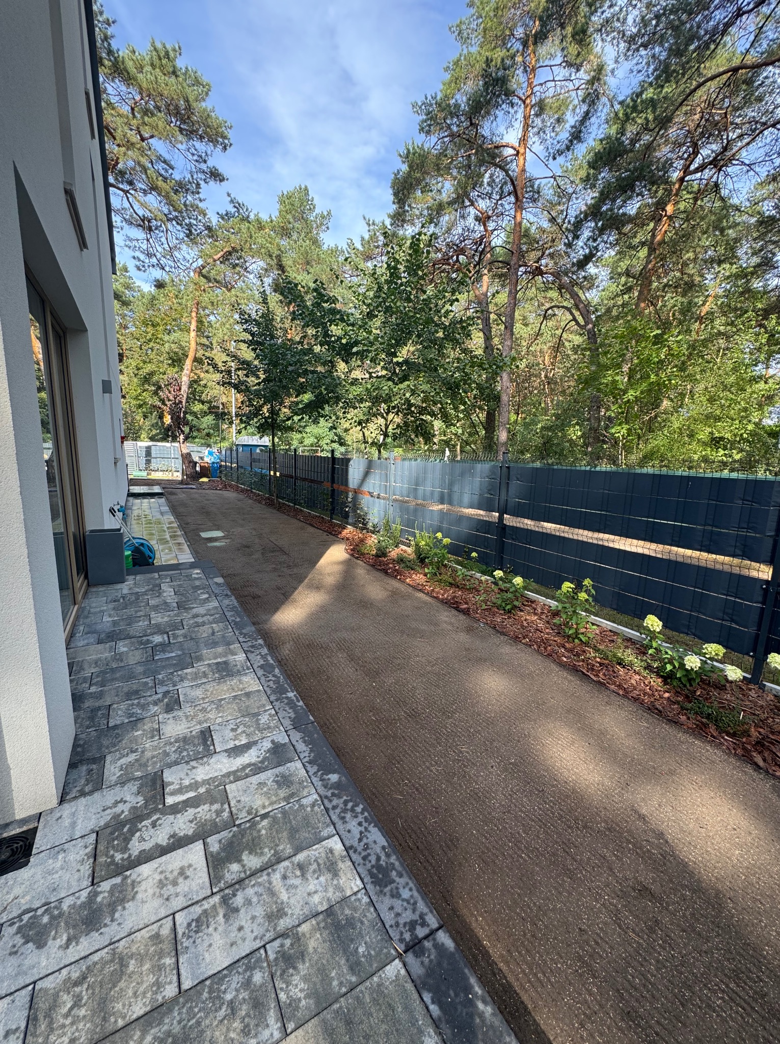 Przygotowany teren pod trawnik z rolki, obrzeża wyłożone korą i posadzone młode krzewy hortensji, widok z boku domu, w tle ogrodzenie panelowe.