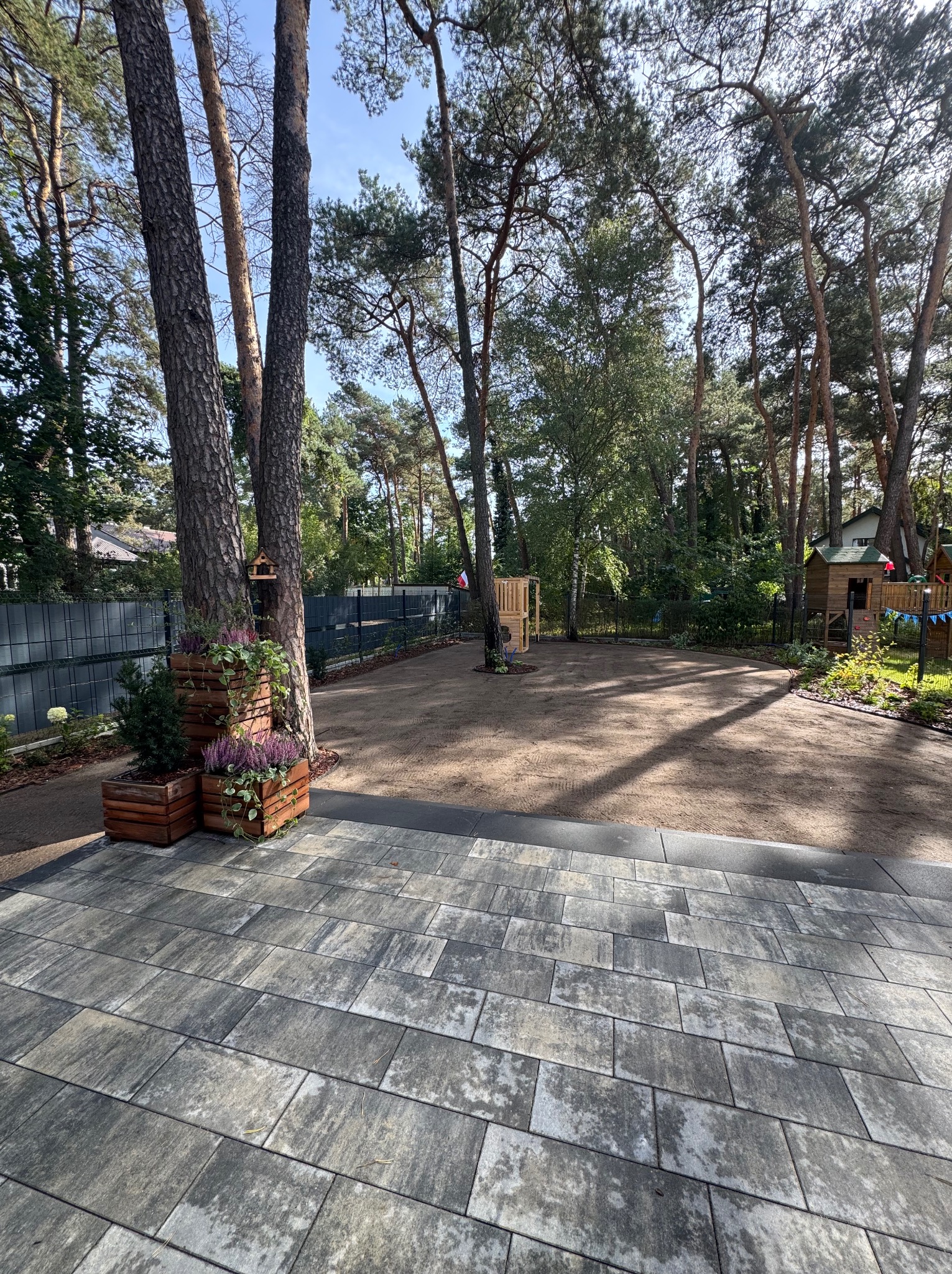 Przygotowany teren pod trawnik z rolki, widok z wybrukowanego tarasu. Na pierwszym planie donice z roślinami, w tle drzewa i elementy placu zabaw.