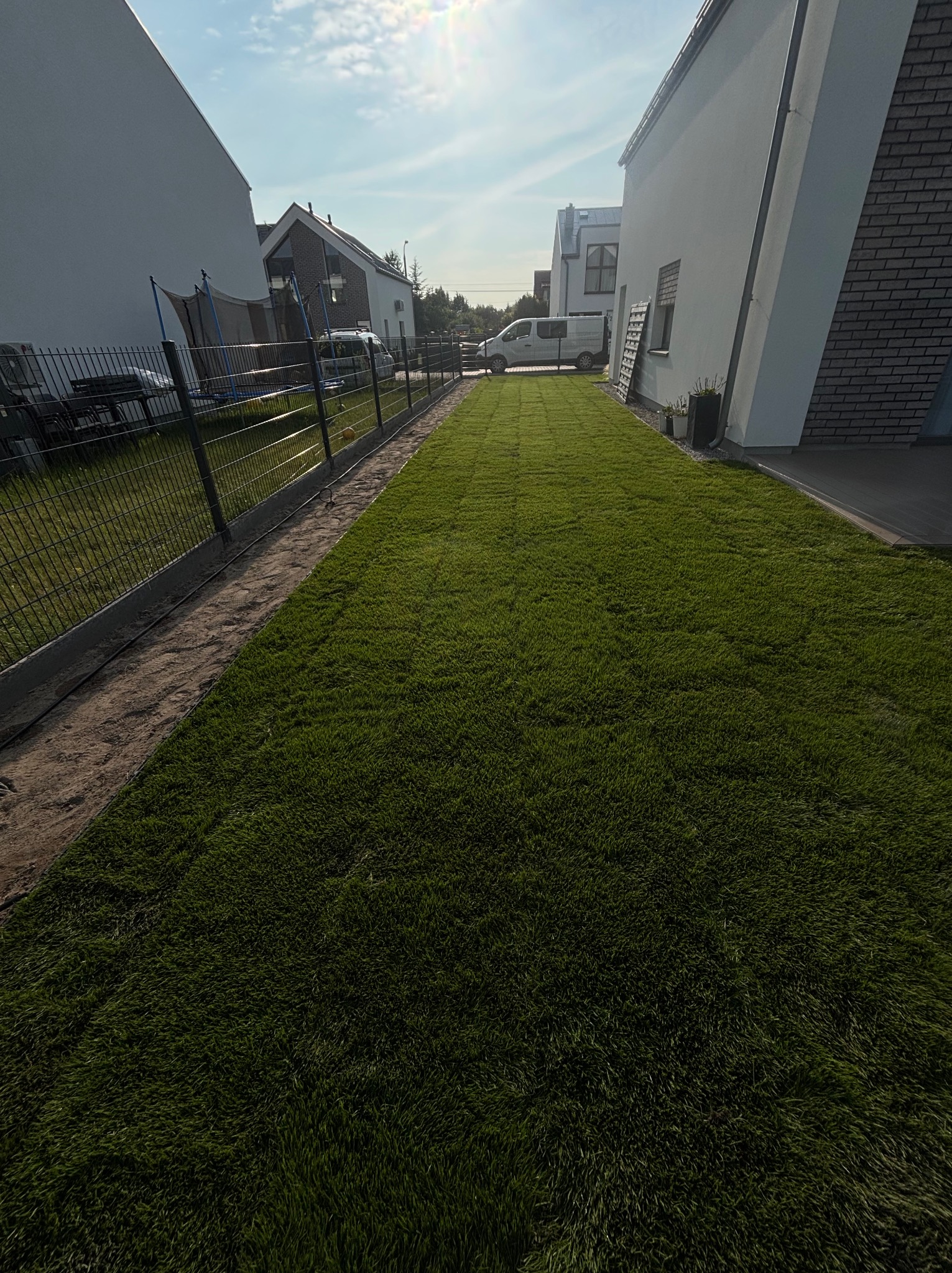 Świeżo ułożona trawa z rolki wzdłuż ogrodzenia nowoczesnego domu, widok z perspektywy trawnika, w tle zaparkowany biały samochód dostawczy.
