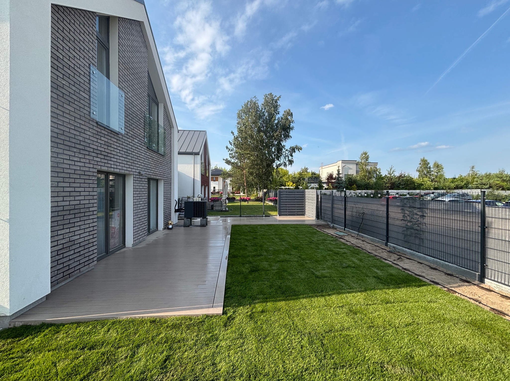 Zadbany trawnik przylegający do nowoczesnego domu z tarasem z desek kompozytowych. Widoczne ogrodzenie panelowe i drzewo w tle, błękitne niebo.