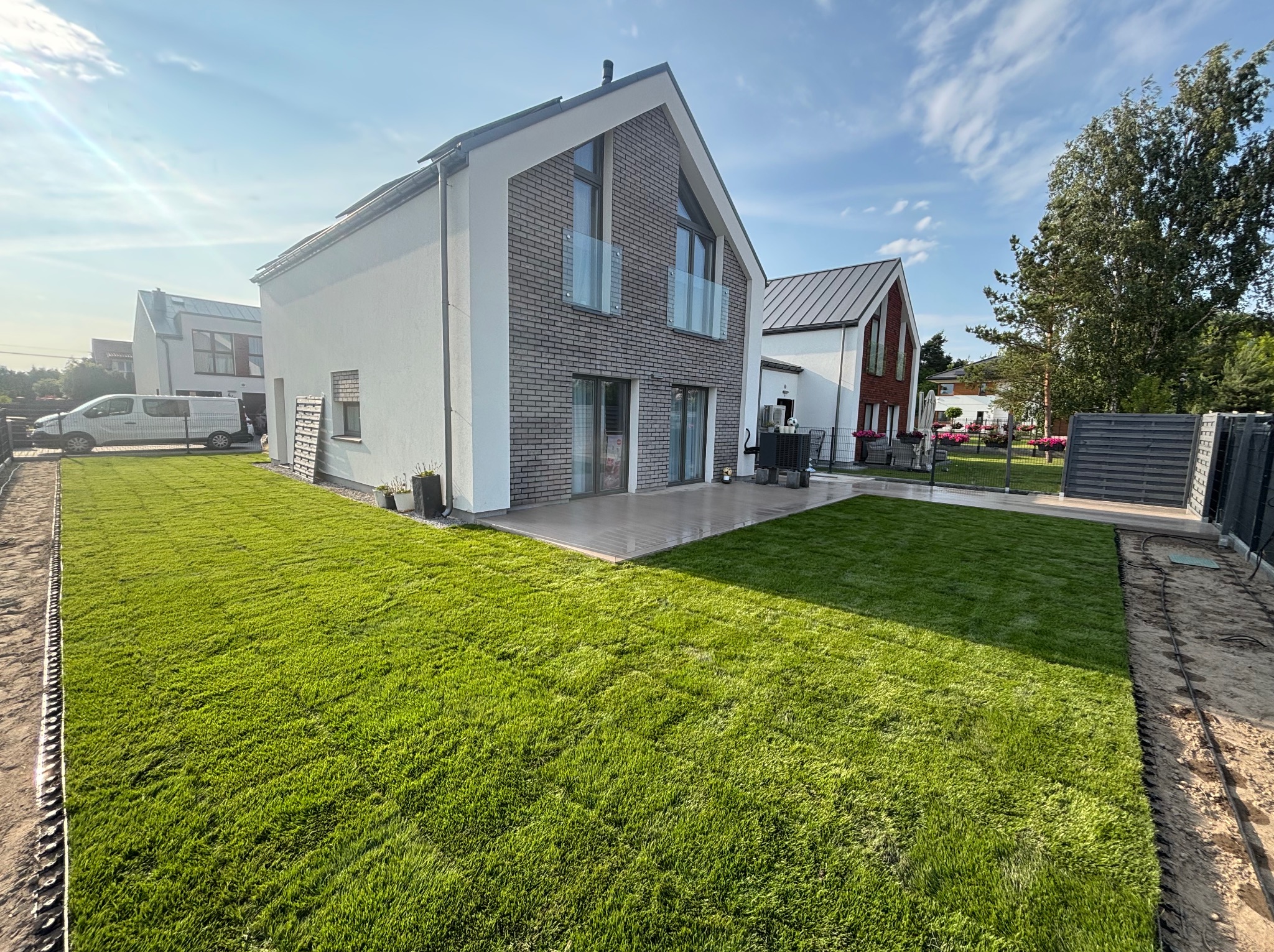 Nowoczesny dom z idealnie ułożoną, soczystą trawą z rolki. Widoczny fragment ogrodu z wyraźną linią łączenia trawy. Słoneczny dzień, błękitne niebo.