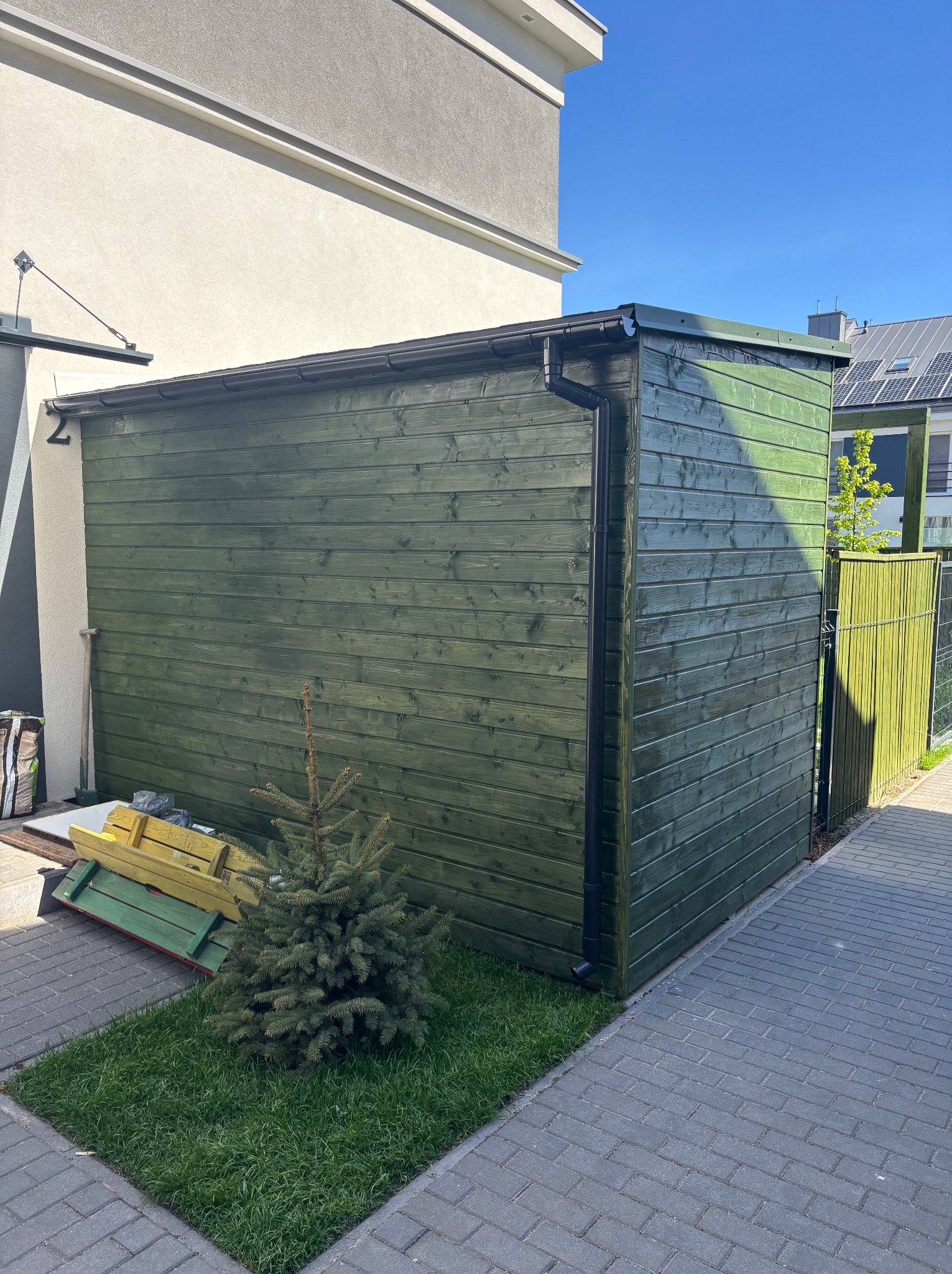 Zielony domek narzędziowy z drewna, obok trawnik z małą choinką. W tle budynek mieszkalny i fragment ogrodzenia. Słoneczny dzień, wyraźne detale drewna.