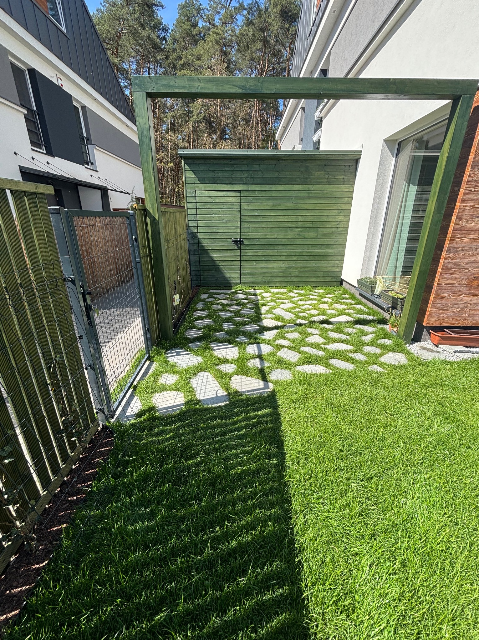 Zadbany trawnik z nieregularnymi kamiennymi płytami prowadzącymi do zielonego domku narzędziowego. Widoczny fragment ogrodzenia i nowoczesna architektura domu w tle.