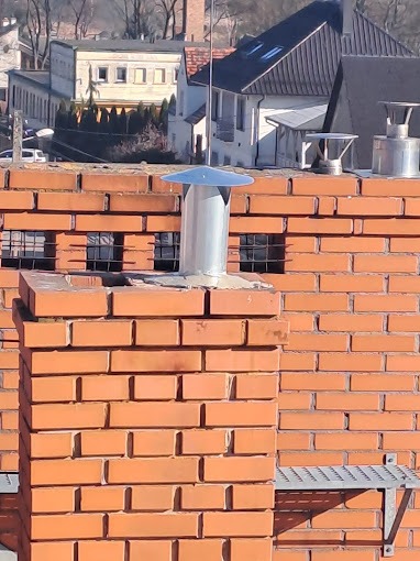 Ceglany komin z metalowym wkładem i daszkiem na dachu budynku, widok z bliska na tle zabudowy miejskiej.