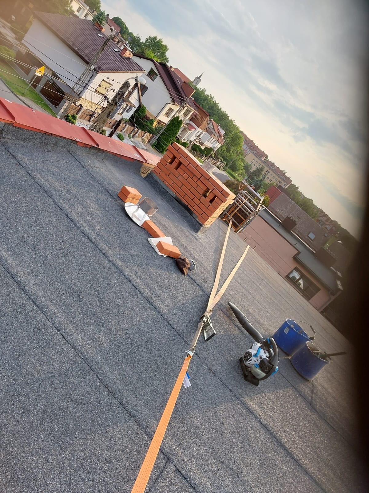 Widok z góry na płaski dach pokryty papą, z przygotowanymi cegłami kominowymi, narzędziami, dmuchawą do liści i zabezpieczeniami, w tle zabudowa miejska.