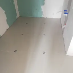 Jasne płytki podłogowe z ciemniejszymi kwadratami w fugach, ściany w surowym stanie, widoczne instalacje hydrauliczne.